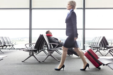 Geschäftsfrau, die einen Koffer an einem ruhenden Geschäftsmann mit Nackenkissen im Abflugbereich eines Flughafens vorbeizieht - CAIF10216