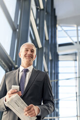 Smiling businessman with cell phone and newspaper in airport - CAIF10265