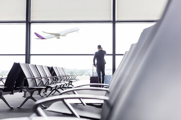 Businessman with suitcase talking on cell phone at airport departure area window - CAIF10277