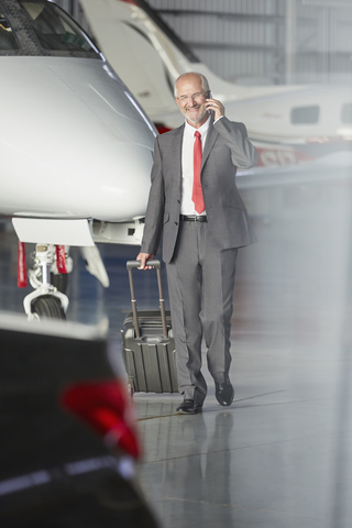 Lächelnder Geschäftsmann, der einen Koffer zieht und mit einem Handy in einem Flugzeughangar telefoniert, lizenzfreies Stockfoto