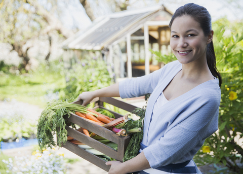 Portrait lächelnde Frau mit frisch geernteter Gemüsekiste im Garten, lizenzfreies Stockfoto