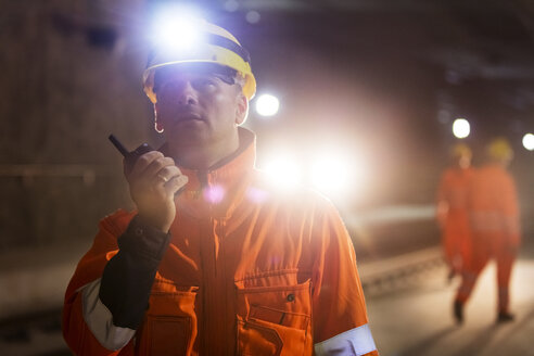 Male foreman with headlamp using walkie-talkie at dark construction site - CAIF10447