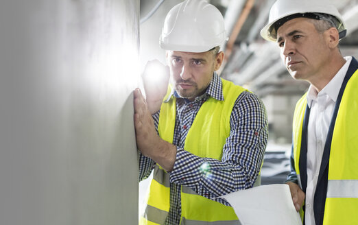 Männlicher Ingenieur mit Taschenlampe, der eine unterirdische Wand auf einer Baustelle untersucht - CAIF10466