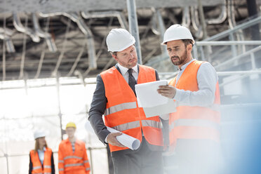 Male engineers with blueprints and clipboard discussing paperwork at construction site - CAIF10470