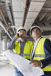 Male engineers with flashlight and blueprints looking up at construction site - CAIF10471