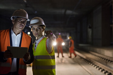 Foreman and engineer with digital tablet talking on dark tracks at underground construction site - CAIF10481