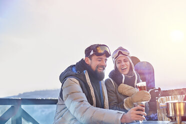 Snowboarder-Paar lacht, trinkt Cocktails auf sonnigem Balkon beim Après-Ski - CAIF10547