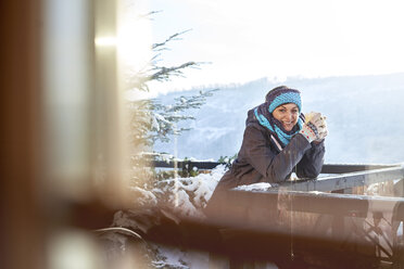 Porträt einer lächelnden Skifahrerin, die auf einem sonnigen Kabinenbalkon einen heißen Kakao trinkt - CAIF10564