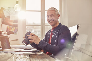 Portrait smiling, confident male photographer with digital camera working at laptop in sunny office - CAIF10581