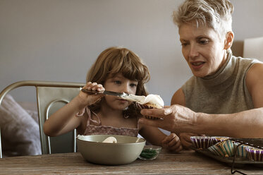 Mädchen trägt Sahne auf einen von der Großmutter gehaltenen Kuchen auf, während sie zu Hause sitzt - CAVF05471