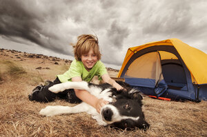 Junge spielt mit Hund, während er bei einem Zelt auf einem Feld sitzt - CAVF05501