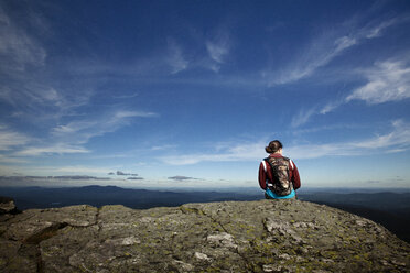 Rear view of woman with backpack sitting on cliff against sky - CAVF05509