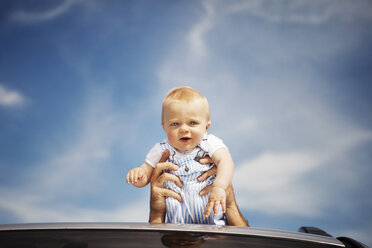 Porträt eines neugierigen kleinen Jungen, der vom Vater über das Autodach gegen den Himmel getragen wird - CAVF05519