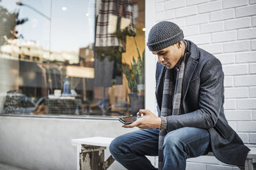 Mann benutzt Mobiltelefon, während er auf einer Bank an der Wand sitzt - CAVF05579