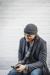 Happy man using mobile phone while sitting against wall - CAVF05580