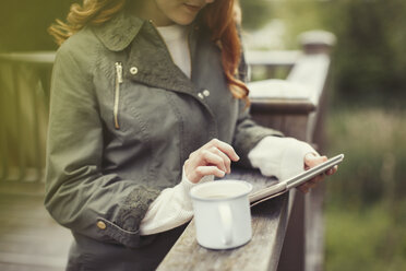 Frau trinkt Kaffee mit digitalem Tablet am Balkongeländer - CAIF10740