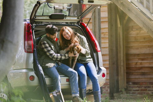 Ehepaar streichelt Hund auf der Rückbank eines Autos vor einer sonnigen Hütte - CAIF10741