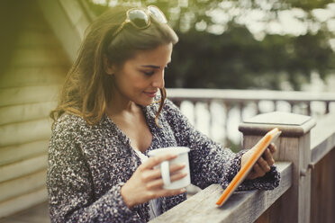 Woman drinking coffee and using digital tablet on balcony - CAIF10748