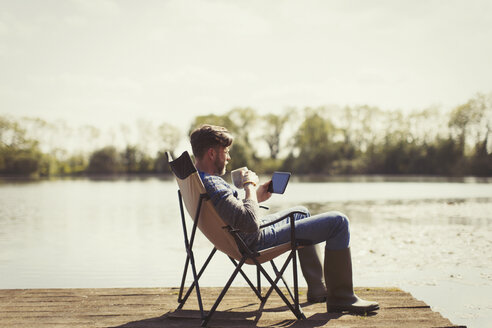 Mann trinkt Kaffee und benutzt ein digitales Tablet auf einem sonnigen Steg am See - CAIF10751