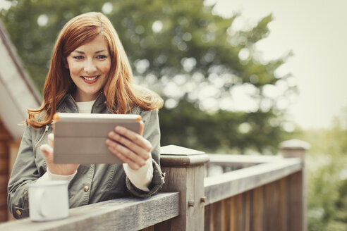Lächelnde Frau mit roten Haaren, die ein digitales Tablet benutzt und am Balkongeländer Kaffee trinkt - CAIF10753