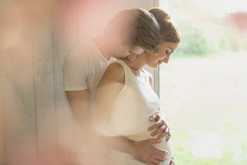 Zärtliches schwangeres Paar, das sich am Fenster umarmt - CAIF10803