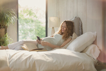 Schwangere Frau liegt entspannt mit Buch und SMS mit Handy im Bett - CAIF10838