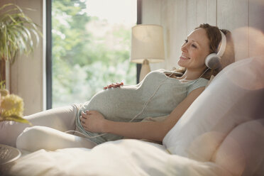 Lächelnde schwangere Frau, die sich beim Musikhören mit Kopfhörern im Bett entspannt - CAIF10841