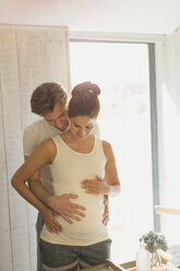 Pregnant couple holding stomach in sunny bathroom - CAIF10844