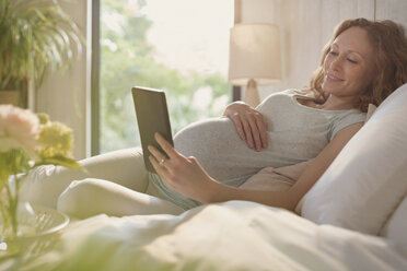 Schwangere Frau entspannt sich mit digitalem Tablet im sonnigen Schlafzimmer - CAIF10864