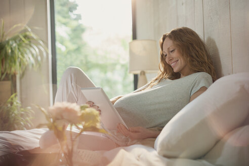 Lächelnde schwangere Frau mit digitalem Tablet auf dem Bett im sonnigen Schlafzimmer - CAIF10878