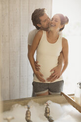 Lachendes liebevolles schwangeres Paar, das sich küsst und ein Schaumbad vorbereitet - CAIF10899