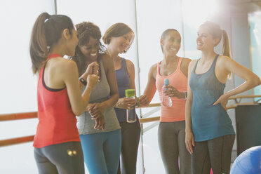 Lächelnde Frauen, die sich in einem sonnigen Fitnessstudio unterhalten und Wasser trinken - CAIF10928