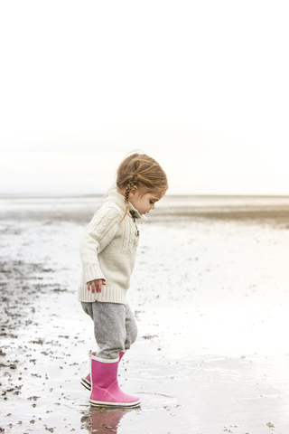 Mädchen in rosa Regenstiefeln spielt im Wasser am Strand, lizenzfreies Stockfoto