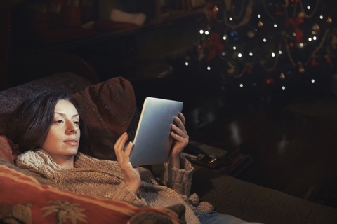 Frau mit digitalem Tablet entspannt auf Sofa im Wohnzimmer in der Nähe von Weihnachtsbaum, lizenzfreies Stockfoto