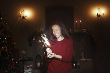 Lächelnde Frau mit Wunderkerze Feuerwerk im dunklen Wohnzimmer mit Weihnachtsbaum - CAIF11142