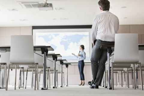 Geschäftsmann beobachtet Geschäftsfrau, die eine Präsentation auf der Projektionsfläche im Konferenzraum hält, lizenzfreies Stockfoto