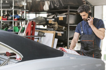 Mechaniker, der mit einem Handy spricht und Autoteile aus einem Katalog in einer Autowerkstatt bestellt - CAIF11237