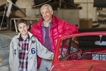 Porträt lächelnder Vater und Sohn neben einem Oldtimer in einer Autowerkstatt - CAIF11258