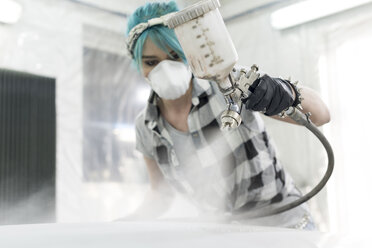 Young woman in protective mask spray painting car in auto body shop - CAIF11261