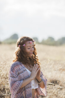 Gelassene Boho-Frau meditiert mit den Händen in der Herzmitte in einem sonnigen ländlichen Feld - CAIF11473