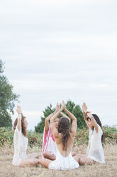 Boho-Frauen meditieren mit über dem Kopf verschränkten Händen in einem Kreis auf einem ländlichen Feld - CAIF11481