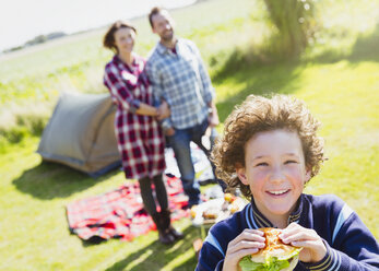 Portrait smiling boy eating hamburger with parents at sunny campsite - CAIF11503