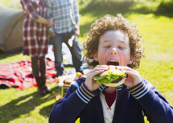 Enthusiastic boy taking large bite of hamburger at sunny campsite - CAIF11513