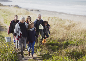 Multi-generation family walking with nets and bucket on sunny grass beach path - CAIF11533