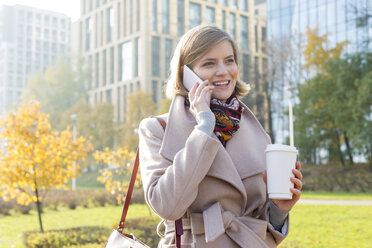 Lächelnde Geschäftsfrau mit Kaffee, die im Stadtpark mit ihrem Handy telefoniert - CAIF11562