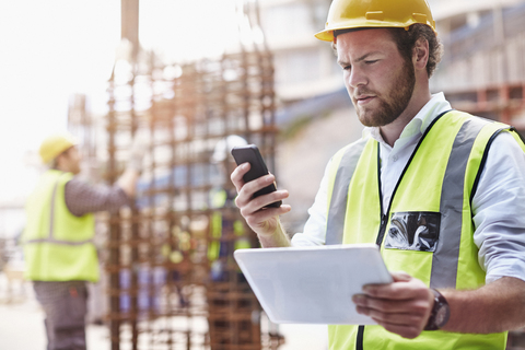 Bauarbeiter mit digitalem Tablet, der auf der Baustelle mit seinem Handy SMS schreibt, lizenzfreies Stockfoto
