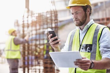 Bauarbeiter mit digitalem Tablet, der auf der Baustelle mit seinem Handy SMS schreibt - CAIF11623