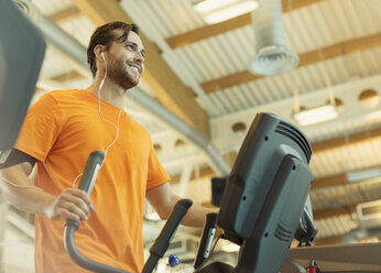 Lächelnder Mann mit Kopfhörern auf einem Ellipsentrainer im Fitnessstudio - CAIF11711