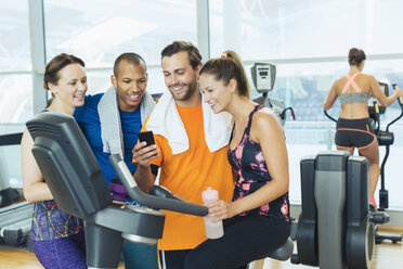 Smiling friends using cell phone at exercise bike in gym - CAIF11785