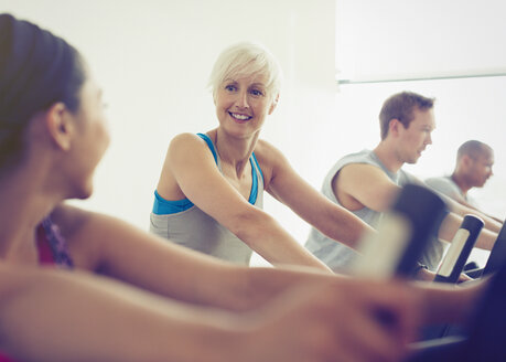 Lächelnde Frauen unterhalten sich auf Heimtrainern im Fitnessstudio - CAIF11789